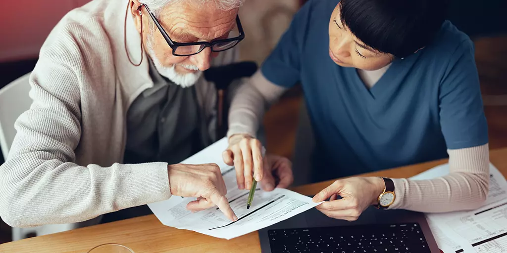 Une professionnelle de la santé aide un homme âgé à remplir des documents administratifs, en pointant des informations sur un formulaire