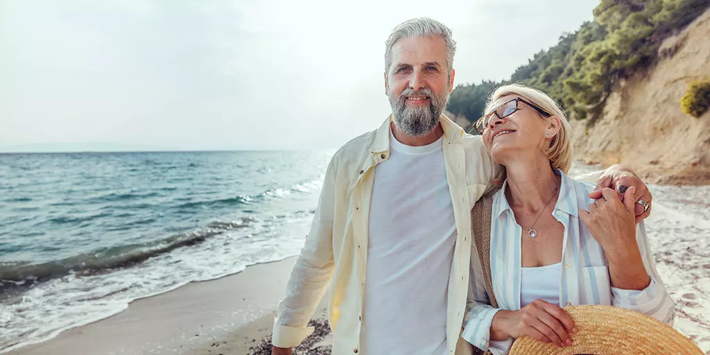 Couple de voyageurs de 65 ans et plus marchant paisiblement sur une plage ensoleillée, souriants et détendus.